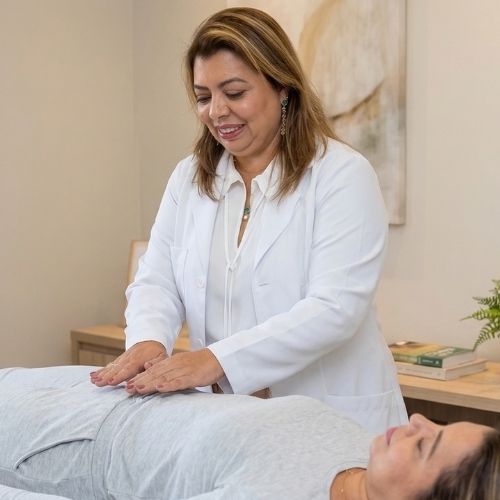 A terapeuta Ana Cecília Beltran, de pele clara, tem cabelos castanho-claros com luzes, abaixo dos ombros, e olhos castanho-claros. Usa duas correntes douradas no pescoço, brincos grandes pendurados e batom rosa. Ela sorri e veste uma blusa branca. Mantém as mãos sobre o abdômen de uma mulher de pele clara, que está deitada, com os olhos fechados. A mulher tem cabelos castanho-claros com luzes, usa batom rosa e uma corrente no pescoço, e veste blusa e calça cinza.