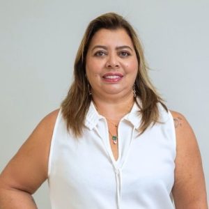 Mulher de pele clara, cabelos loiros abaixo dos ombros, olhos castanhos claros, sorrindo e veste blusa branca.