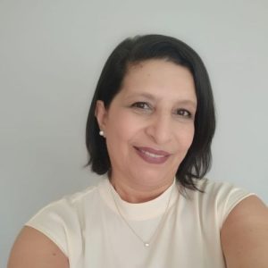 Mulher de pele clara, cabelos curtos castanho-escuros, olhos castanho-escuros, usa brincos, batom marrom e uma corrente no pescoço. Veste uma blusa creme e sorri.