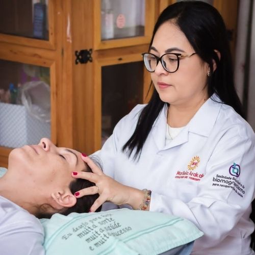 A terapeuta Marileia Arakaki, de pele clara, olhos castanho-escuros e cabelos longos e pretos, usa brincos pequenos, óculos e batom rosa. Ela veste um jaleco branco, com uma camiseta branca por baixo, e uma corrente dourada no pescoço. Usa aliança no dedo anelar, unhas com esmalte vinho e pulseiras com pedras coloridas e cristais. Suas mãos estão sobre a cabeça de uma mulher de pele clara, com cabelos castanho-escuros e olhos fechados, que veste uma camiseta branca e está deitada.