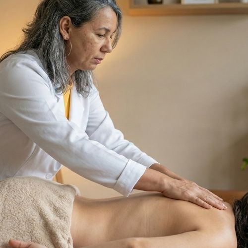 A terapeuta Lurdes Almeida, com cabelos grisalhos abaixo dos ombros, usa batom rosa e brincos de argolas grandes de prata. Ela veste um jaleco branco, com uma blusa amarela por baixo. Suas mãos estão sobre a região torácica de uma mulher deitada de bruços em uma maca, coberta por um lençol bege até a cintura. A mulher tem cabelos grisalhos longos.