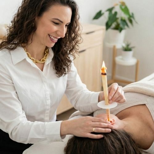 A terapeuta Thaís Meinen, de pele clara, cabelos castanho-escuros, cacheados e longos, veste uma camisa branca e usa batom rosa. Ela usa um colar dourado no pescoço e um anel no dedo anelar da mão direita. Com olhos castanho-escuros e sorri, segura com a mão esquerda um cone hindu aceso próximo ao ouvido esquerdo de uma mulher de pele clara, com cabelos castanho-escuros e longos, que está deitada, veste uma blusa cinza e coberta por um lençol até a altura do peito.