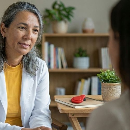 A terapeuta Lu Almeida possui pele clara, olhos castanhos-escuros e longos cabelos grisalhos com luzes. Veste jaleco branco, blusa amarela, brincos de argola e batom rosa. Ela sorri sentada em frente a uma paciente de pele clara, cabelos castanhos-escuros e blusa cinza-clara, que aparece de costas para a câmera.